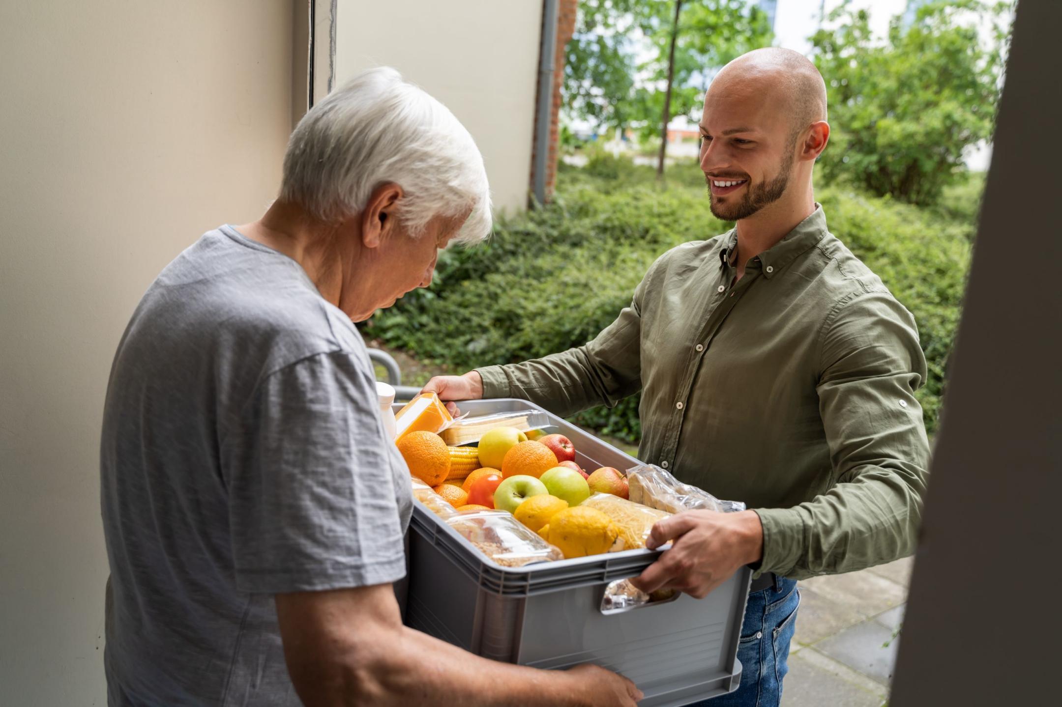 Ein jüngerer Mann bringt einer Senioren Lebensmittel in einer offenen Transportbox an die Haustüre. 