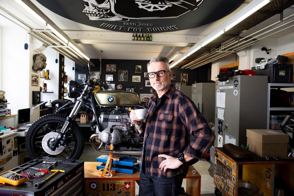 Ein Mitarbeiter in seiner Freizeit mit einer Tasse Kaffee in der Hand in seiner Motorradwerkstatt. Im Hintergrund ein Motorrad auf der Hebebühne, davor liegt Werkzeug.