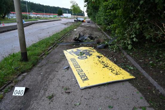 Eine Werbetafel liegt am Boden der Unfallstelle; im Hintergrund das beschädigte Unfallfahrzeug