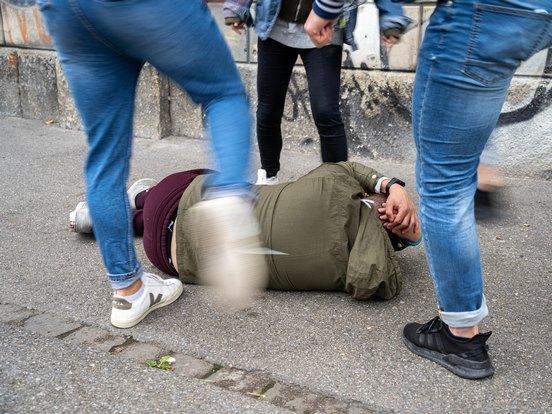 Mehrere Personen traktieren eine am bodenliegende Person mit Fusstritten