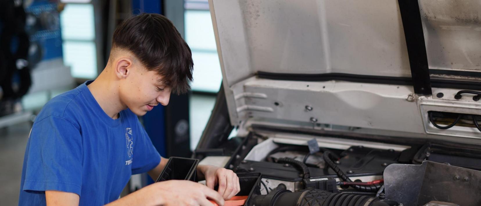 Automobilfachlehrling steht an geöffneter Motorhaube eines Autos und arbeitet.