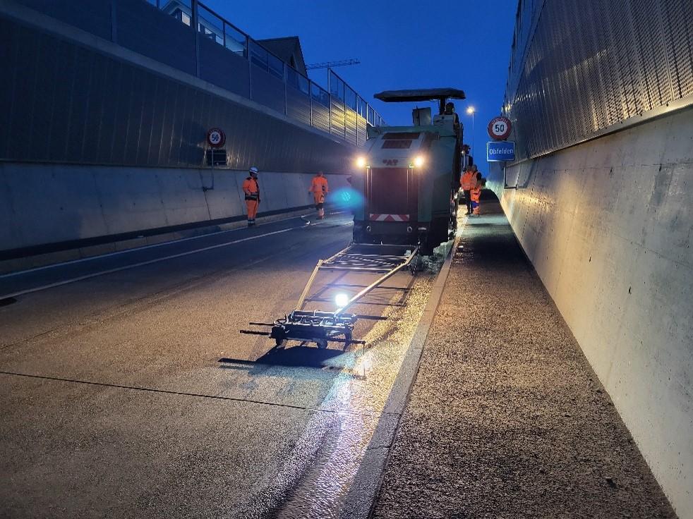 Bauarbeiten bei Nacht auf einer Fahrbahn mit seitlichen Lärmschutzwänden. Eine Maschine zur Oberflächengestaltung ist im Einsatz, umgeben von Arbeitern in orangenen Warnwesten und Helmen. Ein Verkehrsschild weist auf die erlaubte Höchstgeschwindigkeit von 50 km/h hin.