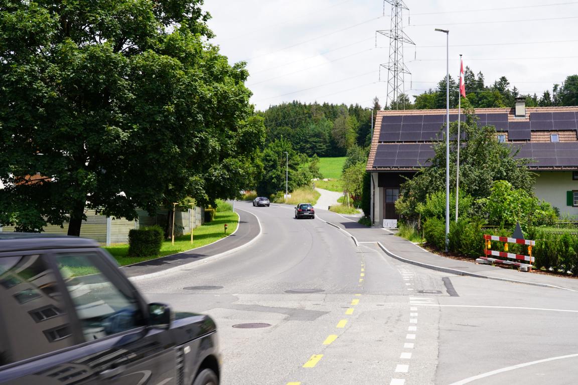Strassenansicht in einem ländlichen Gebiet. Mehrere Autos fahren auf einer kurvigen Strasse leicht bergauf, die von grünen Bäumen umgeben ist. Rechts im Bild befindet sich ein Gebäude mit Solarzellen auf dem Dach und einer Schweizer Flagge. Stromleitungen und ein hoher Strommast sind im Hintergrund sichtbar.