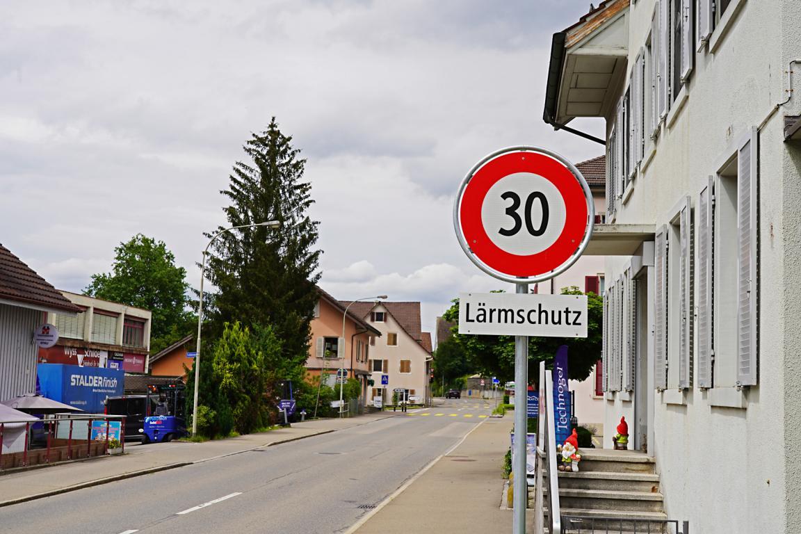 Ein Schild für eine Geschwindigkeitsbegrenzung von 30 km/h zur Lärmschutzmassnahme ist am Strassenrand angebracht. Im Hintergrund sind Wohnhäuser und einige gewerbliche Gebäude zu sehen. Der Himmel ist bewölkt.