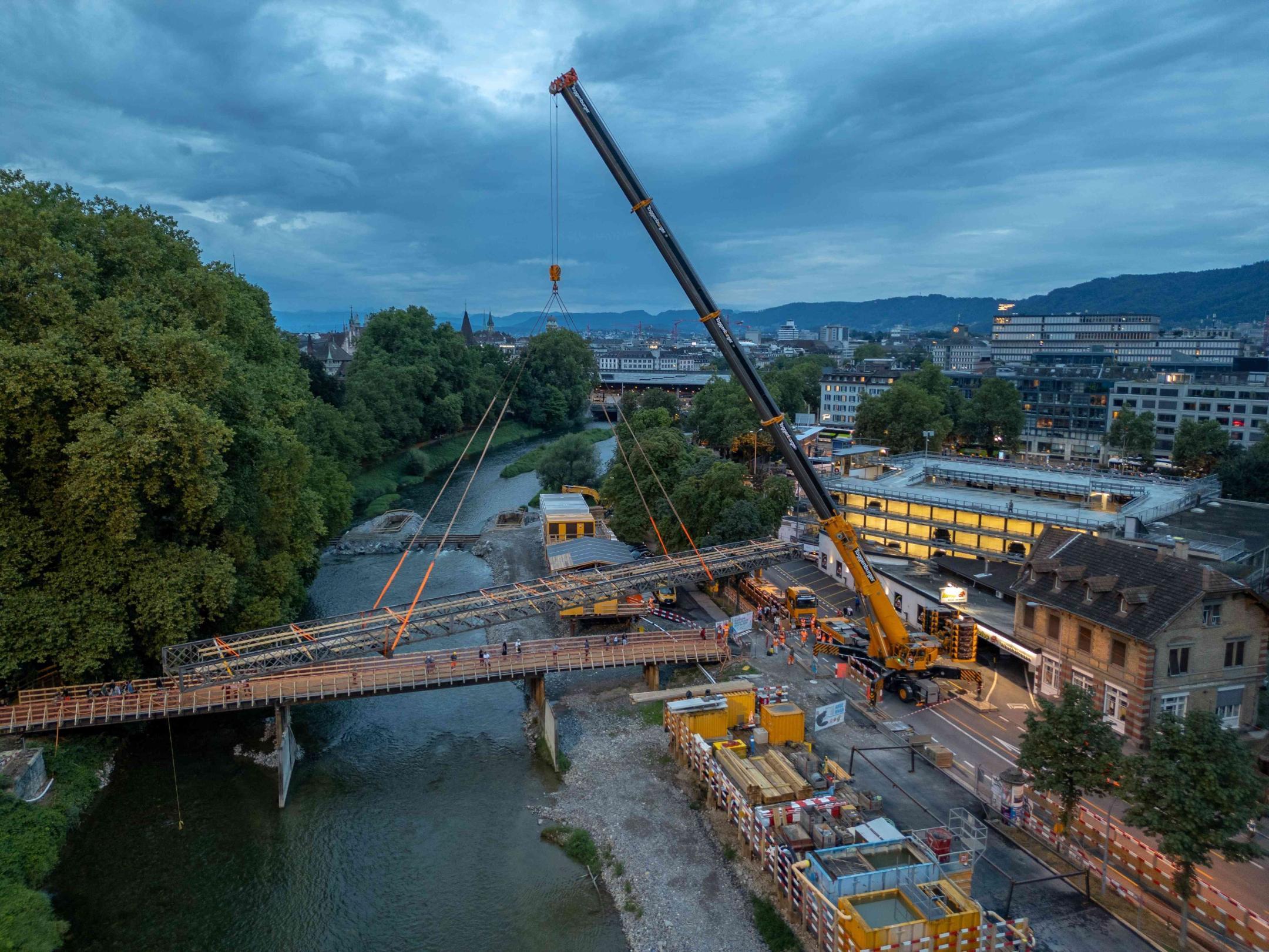 Ein riesiger gelbgrauer Pneukran hebt den Mattensteg am Platzspitz aus der Verankerung. Der Steg schwebt als Ganzes über der Hilfsbrücke über die Sihl. Rundherum die Baustelle für das neue Platzspitzwehr.