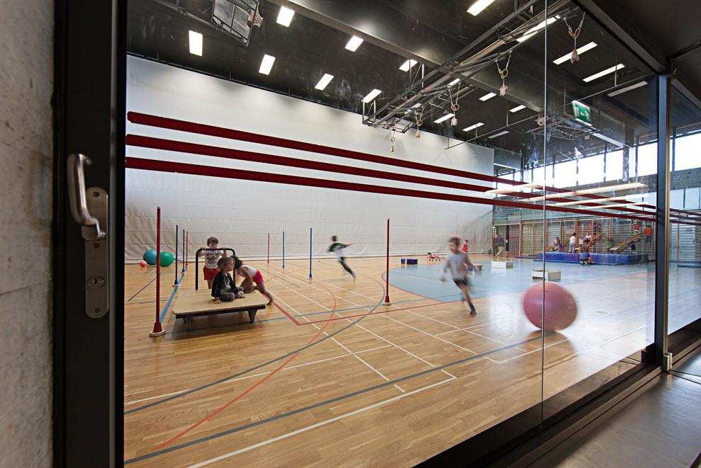 Blick durch eine Scheibe in eine Sporthalle mit Parkett-Boden und spielenden Kindern sowie verschiedenen Sportmaterialien.