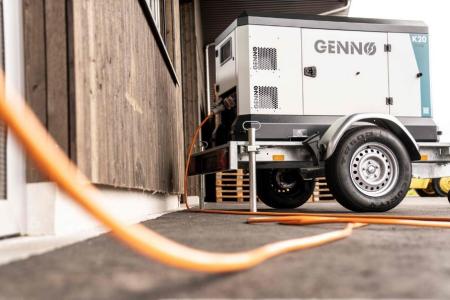 Ein weisses Notstromaggregat mit der Aufschrift Genno auf einem zweiräderigen Anhänger das mit einem langen orangen Kabel verbunden ist.