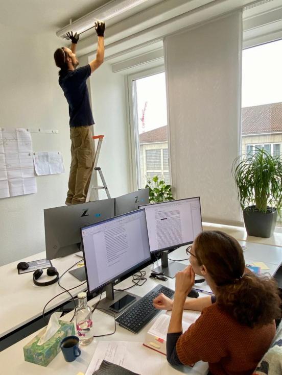 Eine Dame schaut von ihrem Schreibtisch aus zu, wie ein Arbeiter auf einer Leiter die Neonröhre in ihrem Büro ausstauscht. 