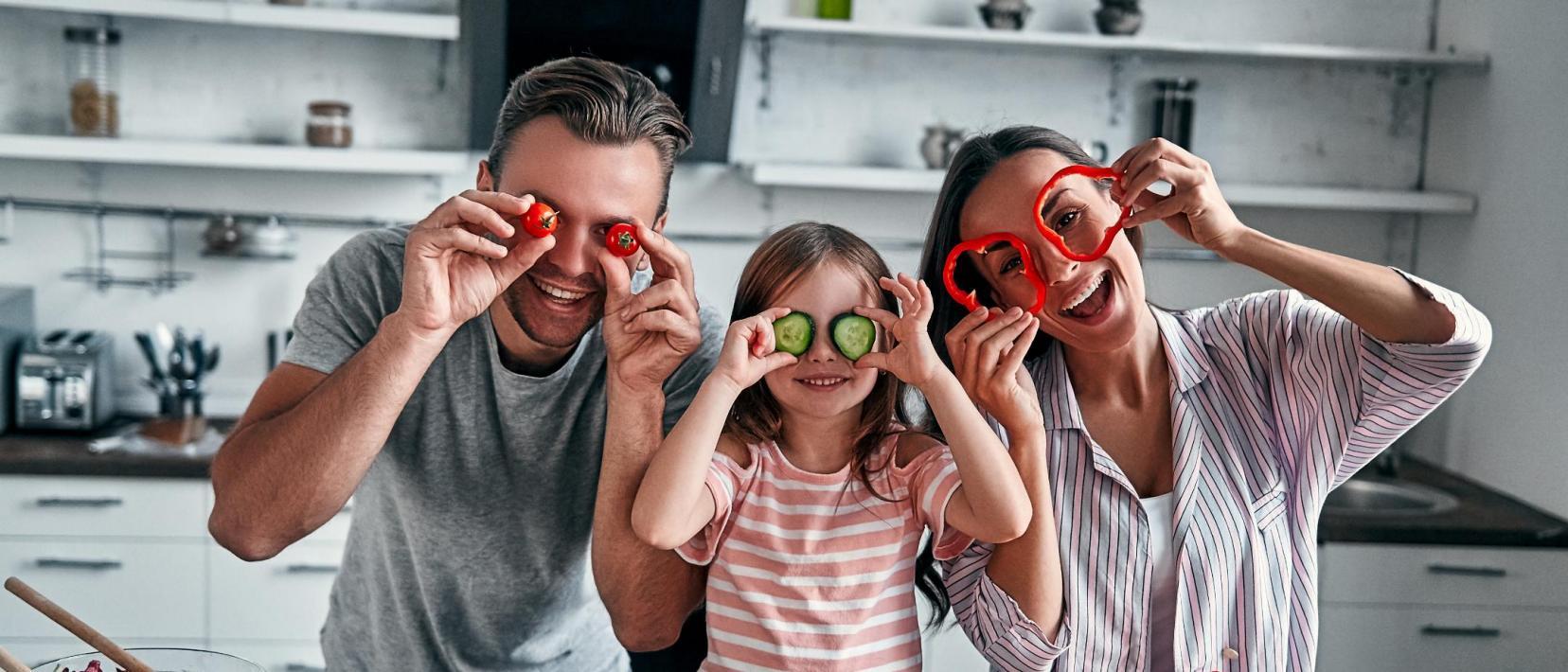 Vater, Mutter und in der Mitte ein Kind stehen an der Küchenzeile und halten lachend Gemüsestücke wie Masken vor ihre Gesichter. Vor ihnen liegen noch mehr Gemüsesorten.
