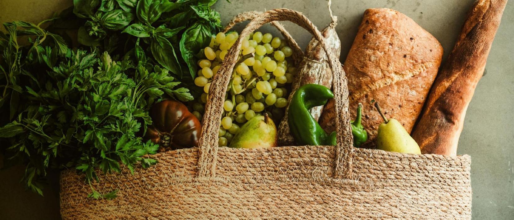 Eine Einkaufstausche aus Naturmaterial, gefüllt mit frischen Kräutern, Gemüse, Früchten und Brot. 