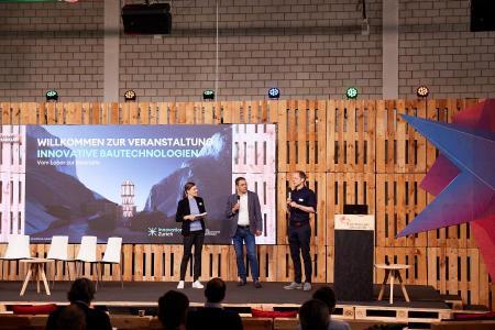 Noémi Besedes, Markus Müller und René Jähne stehen auf der Bühne des Event Hangar im Innovationspark Zürich.