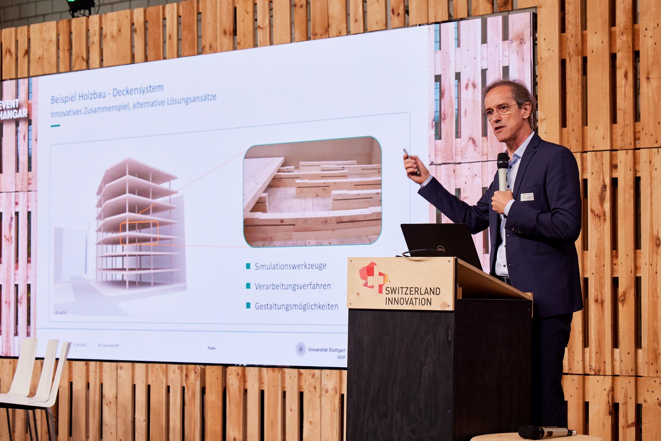 Philip Leistner vom Frauenhofer-Institut für Bauphysik steht auf der Bühne und präsentiert  in einer Power Point Präsentation ein Deckensystem aus Holz. Titel der Präsentation lautet «Beispiel Holzbau - Deckensystem: Innovatives Zusammenspiel, alternative Lösungsansätze».