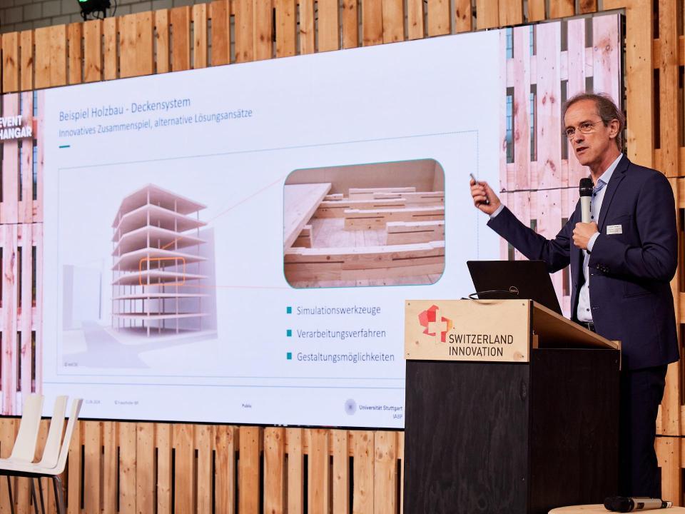 Philip Leistner vom Frauenhofer-Institut für Bauphysik steht auf der Bühne und präsentiert  in einer Power Point Präsentation ein Deckensystem aus Holz. Titel der Präsentation lautet «Beispiel Holzbau - Deckensystem: Innovatives Zusammenspiel, alternative Lösungsansätze».